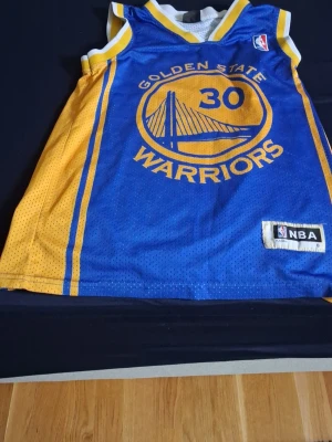 Golden State Warriors Curry tröja - Säljer ett blått och gult basketlinne från Golden State Warriors med nummer 30 och Curry på ryggen. Tröjan har officiell NBA-märkning och är tillverkad i ventilerande meshmaterial, perfekt för basket eller streetwear.
