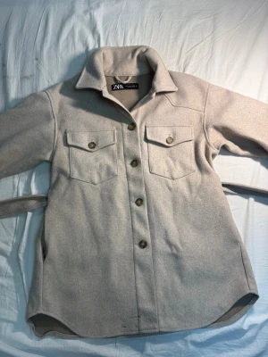 Beige overshirt från Zara - Stilren beige overshirt från Zara med klassisk krage, knäppning framtill och två bröstfickor med lock och knapp. Långärmad modell i mjukt tyg, perfekt för lager på lager. Snyggt avslut nedtill och bälte i midjan för extra passform.