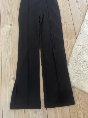 Svarta kostymbyxor med utsvängda ben - Säljer ett par svarta kostymbyxor med utsvängda ben och pressveck. Materialet är slätt och känns dressat, perfekt för en stilren look.
