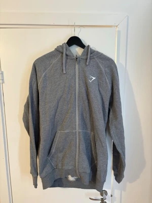 Grå zip hoodie från Gymshark - Säljer en grå zip hoodie från Gymshark med dragkedja framtill, två fickor och justerbar huva med snören. Tillverkad i mjukt sweatshirtmaterial, perfekt för träning eller chill. Gymshark-logga på bröstet ger en sportig look.