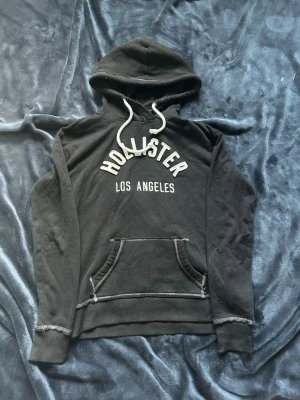 Svart hoodie från Hollister XS - Svart hoodie från Hollister med vit text 'Hollister Los Angeles' på bröstet. Klassisk känguruficka framtill, vita dragsnören i huvan och ribbade muddar. Tillverkad i mjukt bomullsmaterial, perfekt för en avslappnad stil.