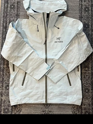 Ljusblå regnjacka från Arc'teryx - Snygg ljusblå regnjacka från Arc'teryx med huva, hel dragkedja och två stora fickor med dragkedja. Jackan har minimalistisk design och är tillverkad i ett vattentätt material som skyddar mot regn och blåst. Perfekt för utomhusaktiviteter.