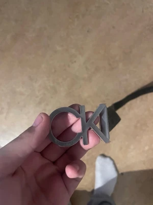 Svart CK bälte med logotypmönster - Snyggt svart bälte från Calvin Klein med silverfärgat CK-spänne. Bältet har ett upprepat CK-mönster över hela remmen , köpt från Zalando orginal , har gjort 1 extra hål i den 