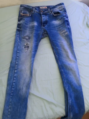 Blå slitna jeans med detaljer från diesel - Säljer ett par blå jeans från Pull&Bear med slitningar och patchade detaljer på låret. Jeansen har klassisk femficksmodell, normal midja och smal passform. Materialet är denim i bomull med stretch för extra komfort. Perfekta för en avslappnad och trendig look.
