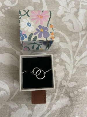 Silverhalsband med två ringar - Snyggt halsband i silverfärg med två sammanlänkade ringar som hänge. Kedjan är tunn och smidig, och halsbandet levereras i en söt ask med blommigt mönster. 