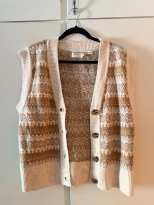 Beige och vit stickad väst från ONLY - Mysig stickad väst från ONLY i beige och vitt med ett klassiskt mönster. Västen har v-ringning, ribbade kanter och stora knappar framtill. Perfekt att slänga över en skjorta eller t-shirt för en trendig lager-på-lager-look.