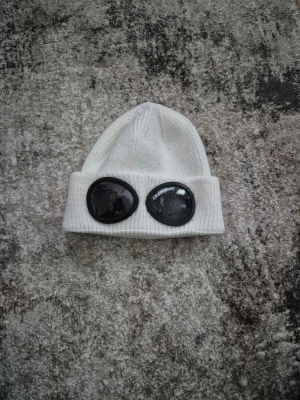 Vit C.P. Company mössa med goggles - Säljer en vit ribbstickad mössa från C.P. Company med de ikoniska svarta goggles framtill. Mössan har uppvikt kant och är perfekt för dig som vill sticka ut med en streetwear-inspirerad look.