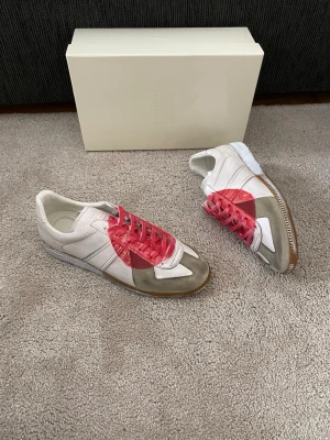 Maison Margiela Japan Gats - Maison Margiela Replica sneakers i vitt läder och mocka med grå och beige detaljer. Skorna har en unik röd färgfläck på sidan och röda skosnören. Klassisk låg silhuett och gummisula. Perfekt för dig som gillar statement sneakers.