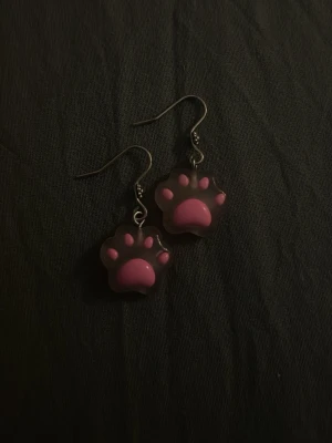 Rosa tassformade örhängen - Säljer ett par söta örhängen formade som tassar i rosa och svart. Örhängena har krokar i metall och själva tassarna är gjorda i plast. Perfekta för dig som gillar gulliga accessoarer med djurtema.