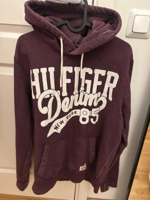 Vinröd Hilfiger hoodie - Snygg vinröd hoodie från Hilfiger Denim med stor vit logga och text på bröstet. Klassisk känguruficka framtill, justerbar huva med vita snören och mjukt material. Perfekt för en avslappnad och trendig look.