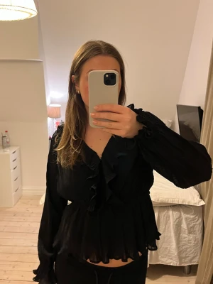 Svart volangblus med långa ärmar - Säljer en svart blus med volangdetaljer längs fram och vid ärmslut. Blusen har långärmade, lätt transparenta ärmar och en v-ringad halsringning. Materialet är tunt och luftigt, perfekt för en trendig look.