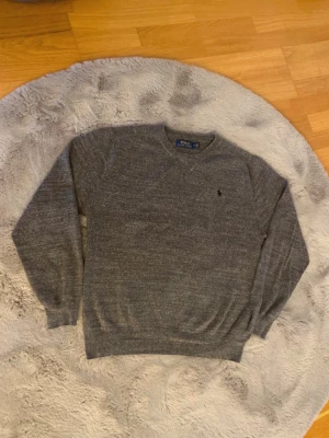 Grå stickad tröja Polo Ralph Lauren - Klassisk grå stickad tröja från Polo Ralph Lauren med rund halsringning och diskret broderad logga på bröstet. Tröjan har långa ärmar och ribbade muddar vid ärmslut och nederkant. Perfekt för lager på lager och enkel att matcha.