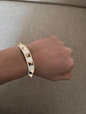Valentino armband - Snyggt vitt armband i läder med guldfärgade pyramidformade nitar längs hela bandet. Justerbart spänne i guldton ger en cool och edgy look. Perfekt accessoar för att lyfta din outfit med lite extra attityd.