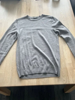 Ljusgrå stickad tröja - En stilren ljusgrå stickad tröja från dressmann med rund halsringning och långa ärmar. Tröjan har ribbade muddar vid ärmslut och nederkant. Perfekt för lager på lager och enkel att matcha med olika outfits.