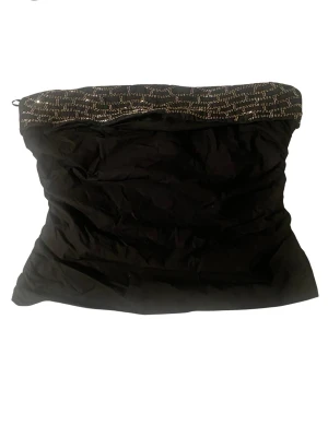 Svart tubtopp med paljettdetalj - Snygg svart tubtopp med glittrande paljetter i guld upptill. Toppen är axelbandslös och har en bandeauformad passning som sitter tight runt överkroppen. Perfekt för dig som vill ha en enkel men ändå festlig look.