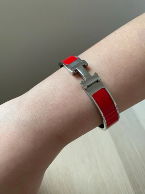 Rött H-armband från H - Snyggt armband från H i silverfärgad metall med röda emaljdetaljer. Armbandet har den ikoniska H-formade låset och en stilren, modern design som verkligen sticker ut. Perfekt accessoar för att lyfta vilken outfit som helst.