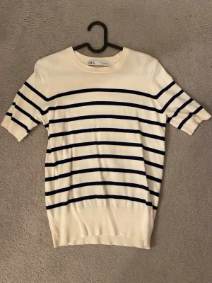 Randig topp från Zara - Vit topp från Zara med marinblå horisontella ränder. Modellen har rund halsringning och korta ärmar. Tillverkad i mjuk bomull, perfekt för en avslappnad och stilren look.