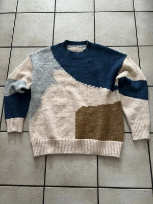 Stickad tröja med grafiskt mönster - Säljer en stickad tröja med coolt grafiskt mönster i beige, blått, grått och brunt. Tröjan har rund hals, ribbade muddar och avslappnad passform. Perfekt för dig som gillar unika plagg med lite retrovibe.