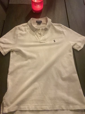 Vit pikétröja från Ralph Lauren - Klassisk vit pikétröja från Ralph Lauren med korta ärmar och krage. Tröjan har knappar vid halsen och den ikoniska broderade loggan på bröstet. Perfekt till jeans eller shorts för en clean och stilren look.