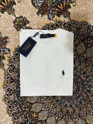 Vit t-shirt från Polo Ralph Lauren - Klassisk vit t-shirt från Polo Ralph Lauren med rund hals och den ikoniska broderade loggan i svart på bröstet. Tillverkad i mjuk bomull för en skön känsla och enkel stil. Perfekt basplagg för alla tillfällen.