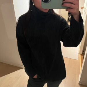 Svart ribbstickad polotröja - Mysig svart ribbstickad polotröja med breda ärmar och hög krage. Tröjan har en avslappnad passform och är perfekt för kalla dagar. Materialet ser ut att vara mjukt och stretchigt, vilket gör den extra bekväm.