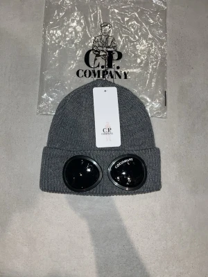 Grå mössa med goggles från C.P. Company - Säljer en grå ribbstickad mössa från C.P. Company med ikoniska svarta goggles framtill. Mössan har uppvikt kant och är tillverkad i mjukt material som ger en skön passform. Perfekt för dig som vill sticka ut med en streetwear-inspirerad look.
