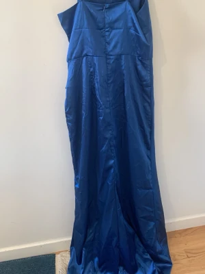 Blå långklänning oneshoulder - Elegant blå långklänning i glansigt material med oneshoulder-design. Klänningen har en figurnära passform upptill och faller snyggt längs kroppen. Perfekt för speciella tillfällen där du vill sticka ut.