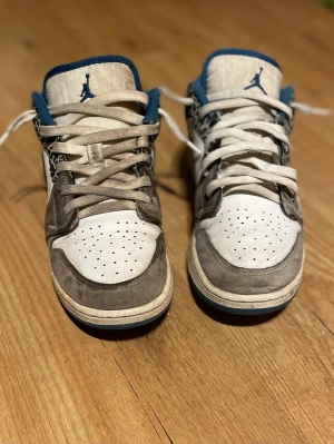 Nike Air Jordan 1 low - Snygga Nike Air Jordan 1 Low med vit och grå ovandel i läder och mocka, blå detaljer på plösen och kragen samt svart Swoosh. Unika elefantmönstrade paneler på sidorna ger extra stilpoäng. Perforerad tåbox för ventilation och klassisk snörning.