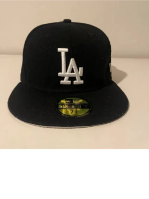 Svart LA New Era 59FIFTY keps - Snygg svart keps från New Era med LA-logga broderad i vitt framtill och klassisk 59FIFTY-sticker på skärmen. Kepsen har rak skärm och New Era-logga på sidan. Perfekt för dig som gillar streetwear och sportig stil.