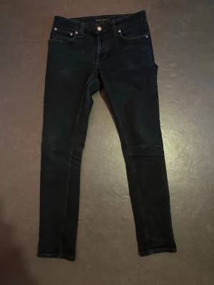 Svarta skinny jeans från Nudie Jeans - Säljer ett par svarta skinny jeans från Nudie Jeans med klassisk femficksmodell och silverfärgade nitar. Jeansen har smal passform och är tillverkade i stretchigt denimtyg för extra komfort. Perfekta för en stilren och modern look.