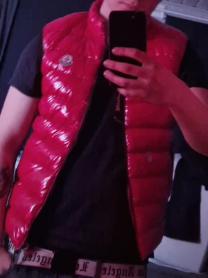 Röd glansig dunväst från Moncler - Snygg röd dunväst från Moncler med glansigt material och svart dragkedja. Västen har två framfickor med dragkedja och Moncler-logga på bröstet. Perfekt för lager på lager och streetwear-vibe.