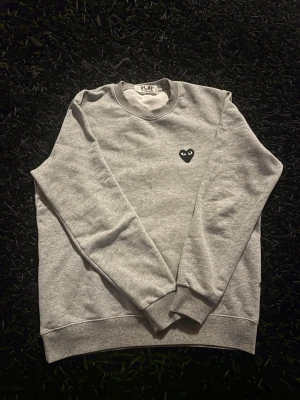 Grå sweatshirt från Comme des Garçons - Säljer en stilren grå sweatshirt från Comme des Garçons Play med det ikoniska svarta hjärtat på bröstet. Tröjan har rund halsringning, ribbade muddar och är tillverkad i mjukt bomullsmaterial. Perfekt för en clean och avslappnad look.