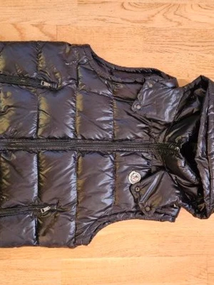 Svart dunväst från Moncler - Snygg svart dunväst från Moncler med glansig finish och avtagbar huva. Västen har två dragkedjeförsedda fickor fram och Moncler-logga på bröstet. Perfekt för lager-på-lager och håller dig varm under kyliga dagar.