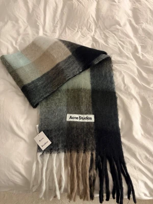 Rutig halsduk från Acne Studios - Mjuk och fluffig halsduk från Acne Studios i rutigt mönster med nyanser av beige, grått, svart och grön. Halsduken har långa fransar i ändarna och en stor Acne Studios-logga. Perfekt accessoar för att lyfta din outfit.