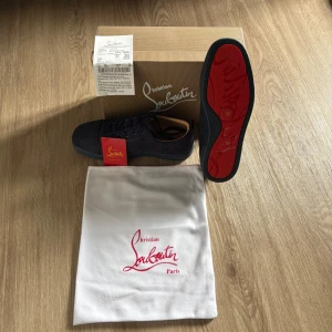 Christian Louboutin marinblå  - Snygga marinblåa sneakers från Christian Louboutin med ikonisk röd sula och guldfärgad logga på insidan. Skorna har ovandel i mocka och skinn, rund tå och klassisk snörning. Perfekt för dig som vill sticka ut med exklusiv stil i sommar. Tveka inte på att höra av dig vid frågor!