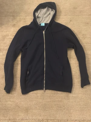 Svart zip hoodie med grått foder - En svart hoodie med dragkedja och grått foder i huvan. Klassisk modell med två fickor framtill och justerbar huva med snören. Perfekt för en chill och avslappnad stil. Tillverkad i mjukt bomullsmaterial.