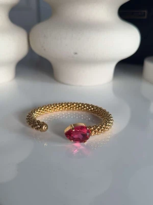 Guldigt armband med rosa sten från Caroline svedbom - Snyggt armband doppad i guld och en stor, droppformad rosa sten framtill. Armbandet är öppet och flexibelt, vilket gör det lätt att ta på och av. Perfekt för dig som vill ha något som sticker ut och känns lyxigt.