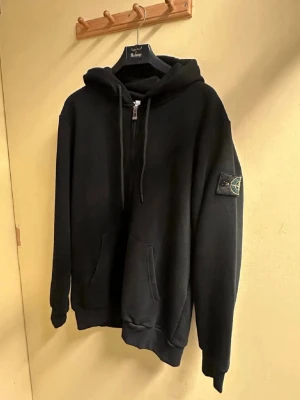 Stone Island, zip hoodie - Svart hoodie från Stone Island med klassisk patch på vänster ärm. Hoodien har dragsko i huvan, magficka och ribbade muddar vid ärmslut och nederkant. Tillverkad i mjukt bomullsmaterial som känns skönt mot huden. Perfekt för en clean och stilren look.