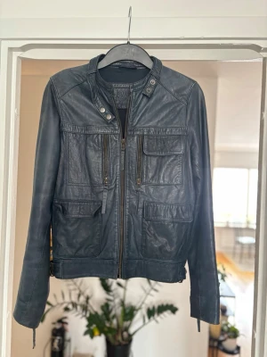 Svart skinnjacka från Hakei - Snygg svart skinnjacka från Hakei med fyra framfickor, dragkedja och coola detaljer vid kragen. Dyr jacka med en hål vid armhålan. Jackan har en rak passform och stilrena sömmar som ger en modern look. Perfekt för dig som vill ha en klassisk men edgy jacka.