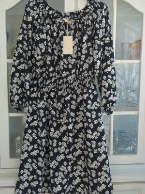 Svart blommig klänning Pernilla Wahlgren - Svart klänning från Pernilla Wahlgren med vitt blommönster. Modellen har lång ärm, resår i midjan och en luftig passform. Perfekt för dig som gillar en feminin och bekväm stil. Tillverkad i mjuk polyester som känns skön mot huden.