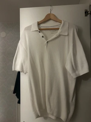 Vit ribbad polotopp med knappar - Säljer en stilren vit polotopp med korta ärmar och ribbad struktur. Toppen har en öppen krage med knappar för en avslappnad look. Perfekt att matcha med jeans för en clean stil till sommaren. Använd 1 gång.
