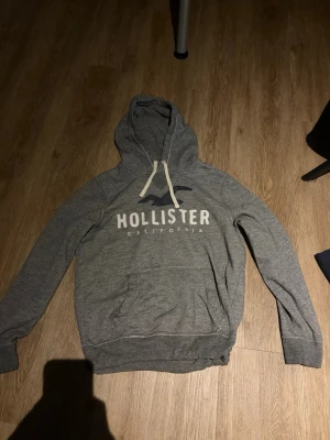 Grå hoodie från Hollister - Snygg grå hoodie från Hollister med stor logga och texten 'Hollister California' på bröstet. Klassisk känguruficka framtill, vita dragsnören i huvan och mjukt material. Perfekt för en avslappnad och trendig look.