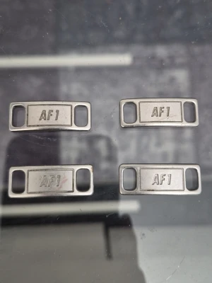 Äkta AF1 skosmycken i metall - Fyra stycken metallfärgade skosmycken med texten 'AF1', perfekta för att piffa upp dina sneakers. Passar till Nike Air Force 1 och ger en snygg detalj till skosnörena. Klassisk rektangulär form med öppningar på sidorna för enkel montering.