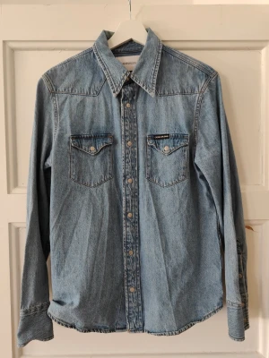 Ljusblå jeansskjorta från Calvin Klein - NYSKICK Snygg ljusblå jeansskjorta från Calvin Klein med klassisk krage, tryckknappar och två bröstfickor med lock. Skjortan har lång ärm och är gjord i bomull för en avslappnad men stilren look. Står XS men passar även S o ksk M