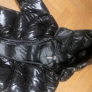 Glansig svart pufferjacka Moncler - Snygg svart pufferjacka från Moncler med glansig finish och hög krage. Jackan har quiltad design, dragkedja framtill och klassisk Moncler-logga på insidan. Perfekt för kalla vinterdagar och riktigt trendig look.