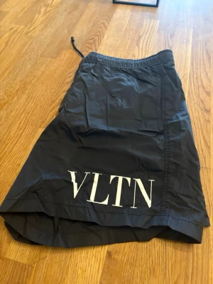 Svarta badbyxor från Valentino - Snygga svarta badbyxor från Valentino med VLTN-logga i vitt på ena benet. Klassisk modell med resår och dragsko i midjan. Tillverkade i lätt och snabbtorkande material, perfekta för stranden eller poolen.