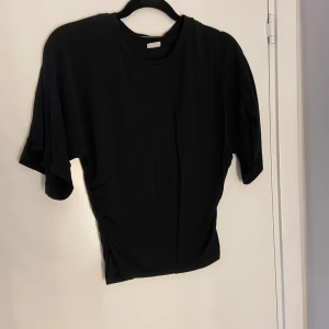 Svart topp med rynkade sidor - Snygg svart topp med korta ärmar och rynkade detaljer längs sidorna som ger en cool och trendig look. Perfekt till jeans eller kjol och enkel att styla upp eller ner. Mjuk och stretchig känsla.