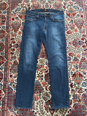Lee jeans - Snygga blå slim fit jeans från Lee med klassisk femficksdesign och subtila slitningar framtill. Jeansen är lite slitna vid föllarna men annars är de i bra skick. Vid andra frågor eller funderingar tveka inte på att höra av dig!