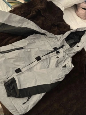 Grå vindjacka från 157.FUNK - Snygg grå vindjacka från 157.FUNK med svarta detaljer på ärmar och huva. Jackan har dragkedja framtill, justerbar huva och flera praktiska fickor. Perfekt för blåsiga dagar och outdoor-aktiviteter. Materialet är lätt och vattenavvisande. Även reflex på hela jackan, nästan aldrig använd.