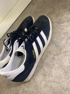 Adidas Gazelle svarta sneakers - ‼️skorna tvättas innan postningen ‼️Klassiska Adidas Gazelle sneakers i svart mocka med vita detaljer och de ikoniska tre ränderna på sidan. Låg modell med svart snörning och vit sula. Guldfärgad Gazelle-text på sidan och Adidas-logga på hälen och plösen.